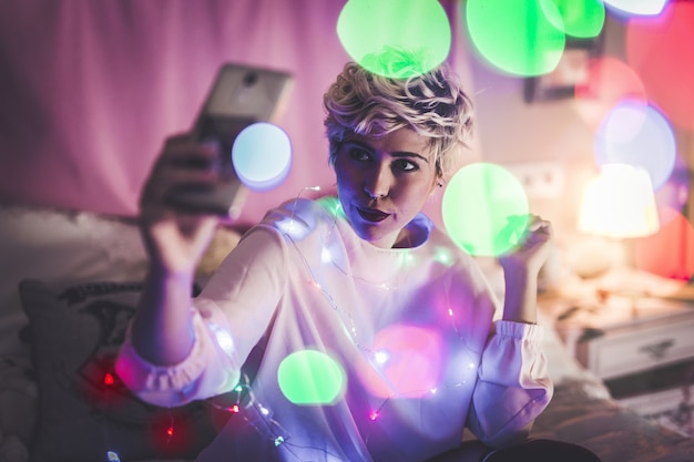 Mujer joven tomando un selfie en su habitación con luces de Navidad