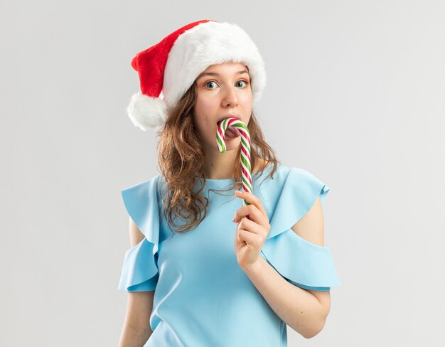 Mujer joven en top azul y gorro de Papá Noel con bastón de caramelo feliz y alegre que va a degustarlo