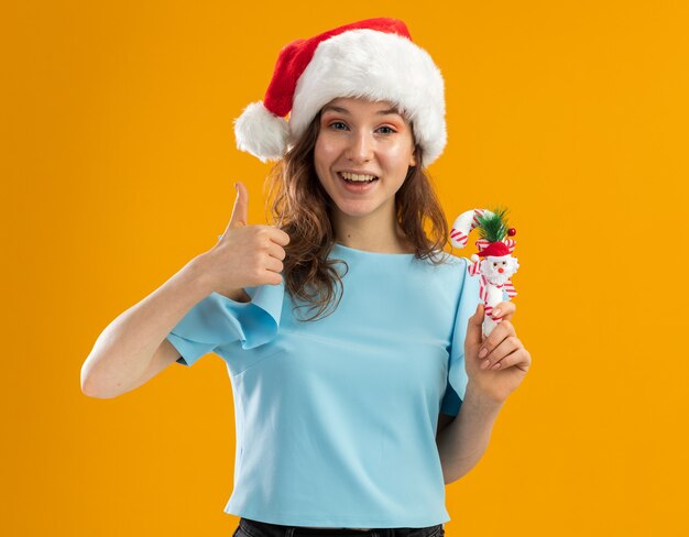 Mujer joven en top azul y gorro de Papá Noel con bastón de caramelo de Navidad mirando sonriendo alegremente mostrando los pulgares para arriba