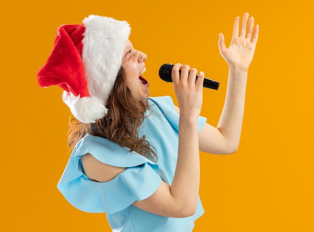 Foto gratuita mujer joven en top azul y gorro de papá noel gritando al micrófono emocionado loco emocional con el brazo levantado