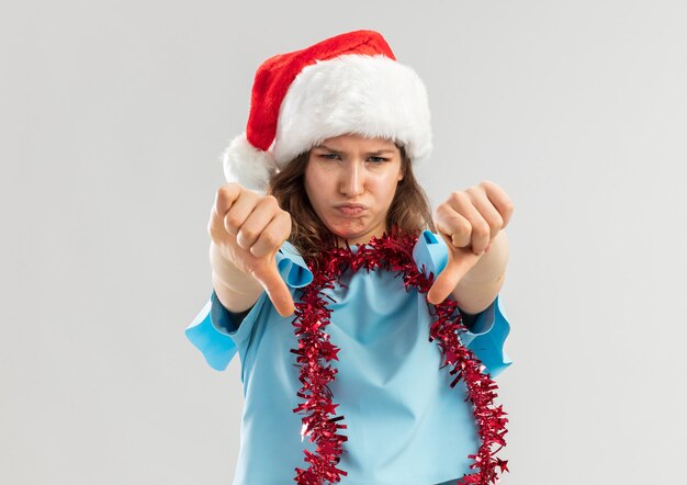 Mujer joven en top azul y gorro de Papá Noel con oropel alrededor de su cuello mirando disgustado mostrando Thumbs dwon