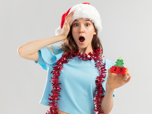 Mujer joven en top azul y gorro de Papá Noel con oropel alrededor de su cuello sosteniendo cubos de juguete con fecha de Navidad mirando asombrado con la mano en la cabeza