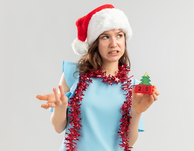 Mujer joven en top azul y gorro de Papá Noel con oropel alrededor de su cuello sosteniendo cubos de juguete con fecha de Navidad mirando a un lado confundido y disgustado con el brazo extendido