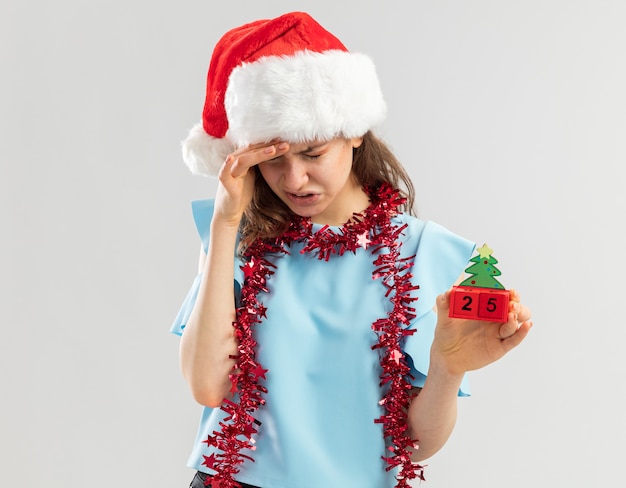 Mujer joven en top azul y gorro de Papá Noel con oropel alrededor de su cuello sosteniendo cubos de juguete con fecha de Navidad tocando su cabeza con aspecto estresado