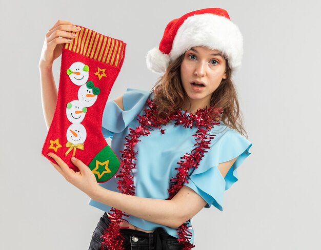 Mujer joven en top azul y gorro de Papá Noel con oropel alrededor de su cuello sosteniendo una media de Navidad sorprendida de pie sobre la pared blanca