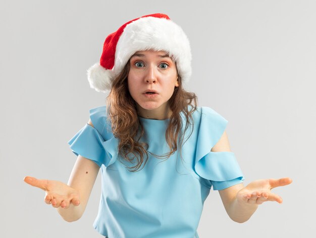 Mujer joven en top azul y gorro de Papá Noel que parece confundido y muy ansioso levantando las manos con indignación y disgusto