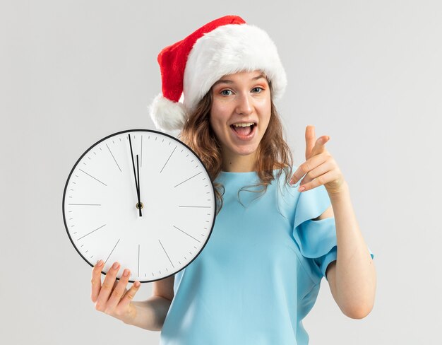 Mujer joven en top azul y gorro de Papá Noel con reloj de pared apuntando con el dedo índice feliz y positivo