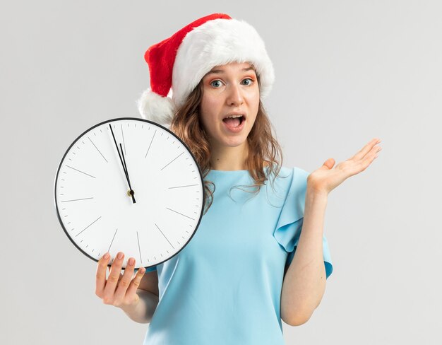 Mujer joven en top azul y gorro de Papá Noel con reloj de pared mirando sorprendido y feliz con el brazo levantado