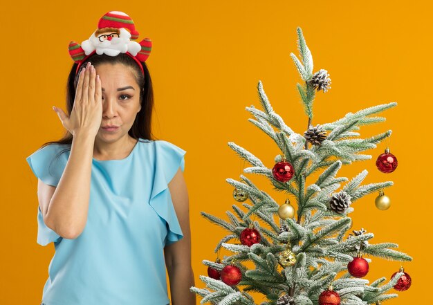 Mujer joven en top azul con gracioso borde navideño en la cabeza mirando a la cámara con rostro serio cubriendo un ojo con la mano de pie junto a un árbol de navidad sobre fondo naranja