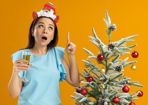 Mujer joven en top azul con gracioso borde navideño en la cabeza sosteniendo una copa de champán apuntando hacia arriba siendo surprsied de pie junto a un árbol de navidad sobre una pared naranja