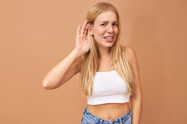 Mujer joven en top blanco y jeans manteniendo la mano en la oreja, escuchando atentamente