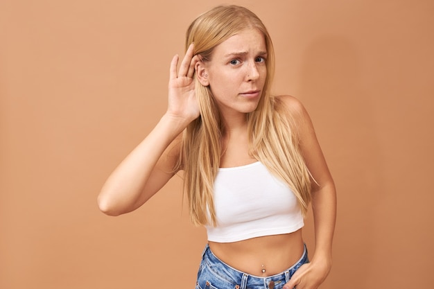 Foto gratuita mujer joven en top blanco y jeans manteniendo la mano en la oreja, escuchando atentamente