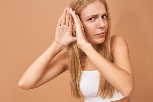 Mujer joven en top blanco y jeans manteniendo la mano en la oreja, escuchando atentamente