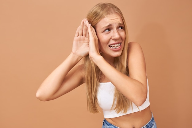 Mujer joven en top blanco y jeans manteniendo la mano en la oreja, escuchando atentamente