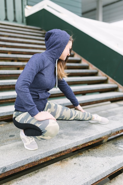 Mujer joven en top con capucha que estira su pierna en escalera