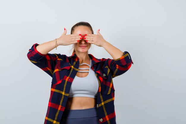 Mujer joven en top corto, camisa a cuadros, pantalones con las manos en los ojos y un aspecto lindo, vista frontal.