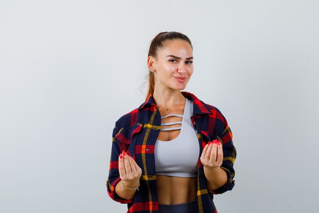 Mujer joven en top corto, camisa a cuadros, pantalones mostrando gesto italiano y mirando encantada, vista frontal.