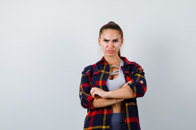 Mujer joven en top corto, camisa a cuadros, pantalones de pie con los brazos cruzados y mirando confiado, vista frontal.