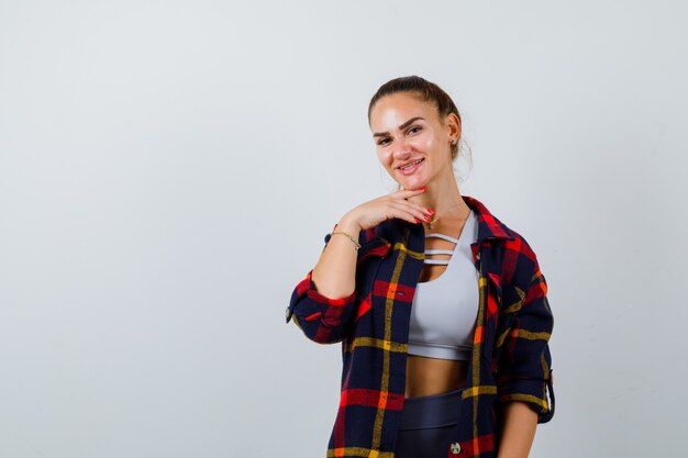 Mujer joven en top corto, camisa a cuadros, pantalones posando mientras está de pie y mirando alegre, vista frontal.