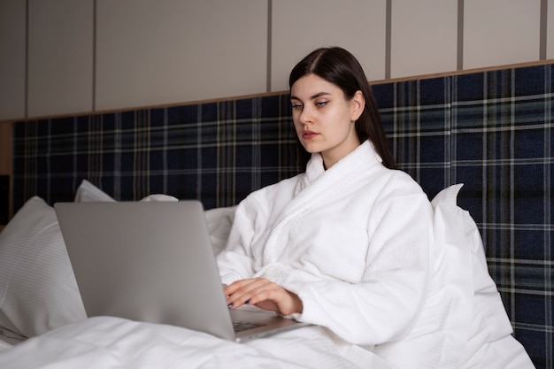 Foto gratuita mujer joven trabajando en su computadora portátil mientras está sentada en la cama en una habitación de hotel