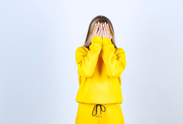 Mujer joven en traje amarillo que cubre su rostro sobre la pared blanca