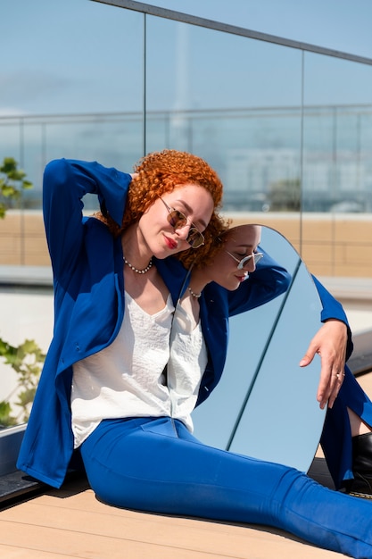 Mujer joven en traje azul posando con espejo al aire libre