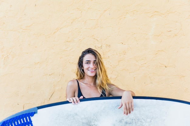 Mujer joven en traje de baño con tabla de surf en la pared amarilla
