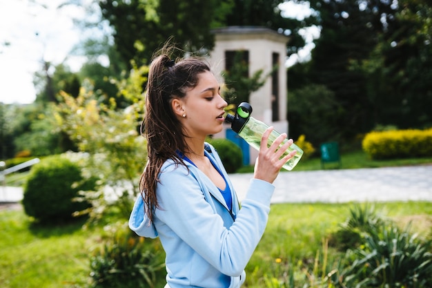 Mujer joven en traje deportivo bebe agua de una botella después de la gimnasia al aire libre en el verano