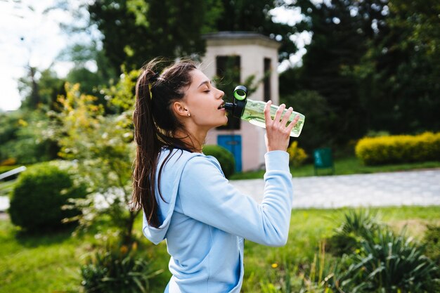 Mujer joven en traje deportivo bebe agua de una botella después de la gimnasia al aire libre en el verano