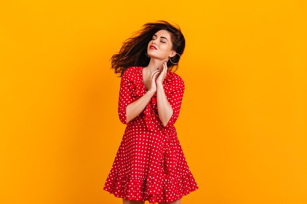 Mujer joven en traje de lunares rojos con sonrisa y ojos cerrados posando en la pared amarilla.