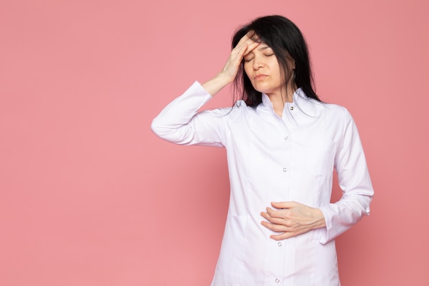 mujer joven en traje médico blanco con un fuerte dolor de cabeza en rosa