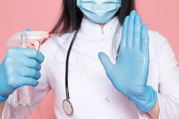mujer joven en traje médico blanco guantes azules máscara protectora azul con estetoscopio con spray desinfectante en rosa