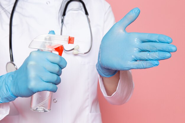 mujer joven en traje médico blanco guantes azules máscara protectora azul con estetoscopio con spray desinfectante en rosa