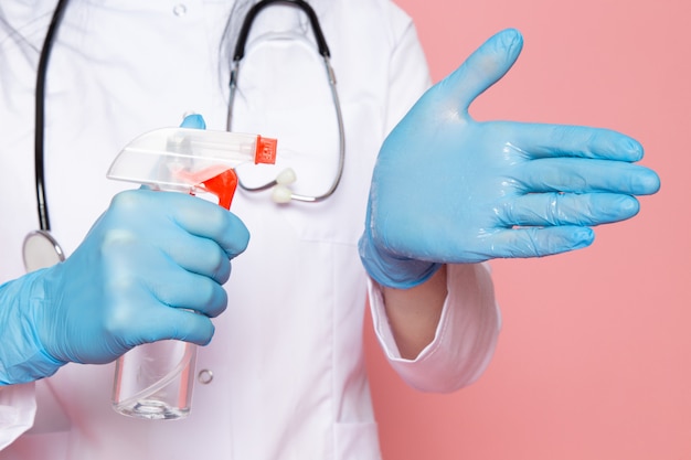 mujer joven en traje médico blanco guantes azules máscara protectora azul con estetoscopio con spray desinfectante en rosa