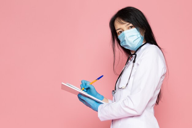 mujer joven en traje médico blanco guantes azules máscara protectora azul con estetoscopio tomando notas en rosa