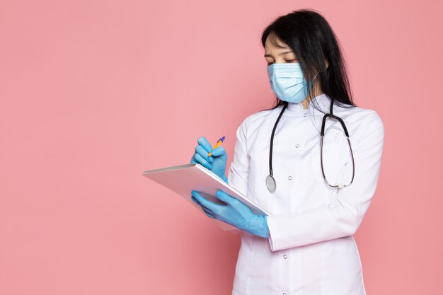 mujer joven en traje médico blanco guantes azules máscara protectora azul con estetoscopio tomando notas en rosa