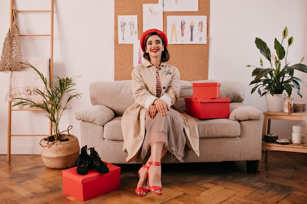 Mujer joven en traje de otoño de moda posa en la acogedora habitación. Una mujer bonita en ropa de moda y tacones rojos está sentada en un sofá beige junto a cajas rojas.