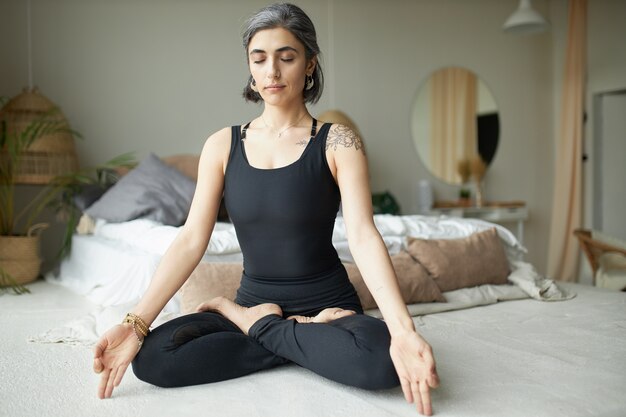 Mujer joven tranquila y pacífica con cabello gris, anillo en la nariz y tatuaje manteniendo los ojos cerrados mientras practica la meditación después del yoga