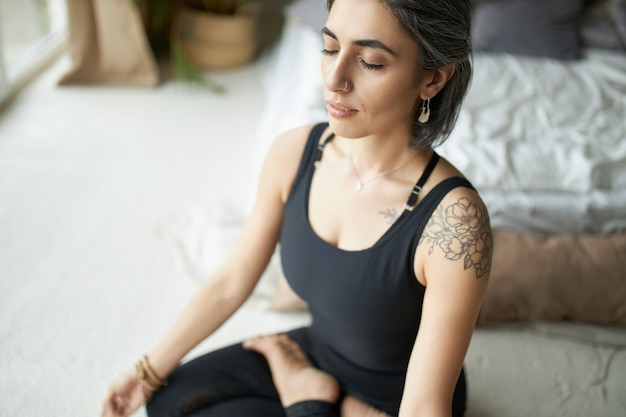 Mujer joven tranquila y pacífica con cabello gris, anillo en la nariz y tatuaje manteniendo los ojos cerrados mientras practica la meditación después del yoga