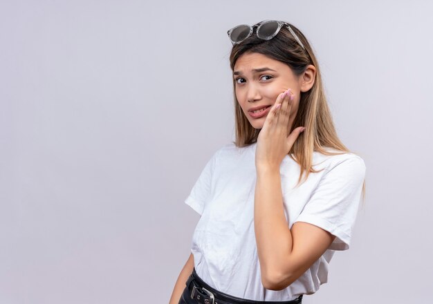 Una mujer joven triste en camiseta blanca con gafas de sol manteniendo la mano en la mejilla y teniendo dolor mientras mira en una pared blanca