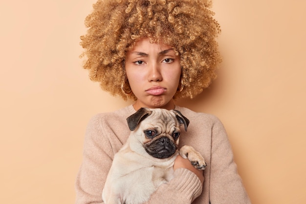 Una mujer joven triste y molesta posa con un perro pug en las manos tiene mal humor ya que su mascota va a tener una visita al veterinario. Los labios se sienten decepcionados vestidos casualmente aislados sobre fondo beige.