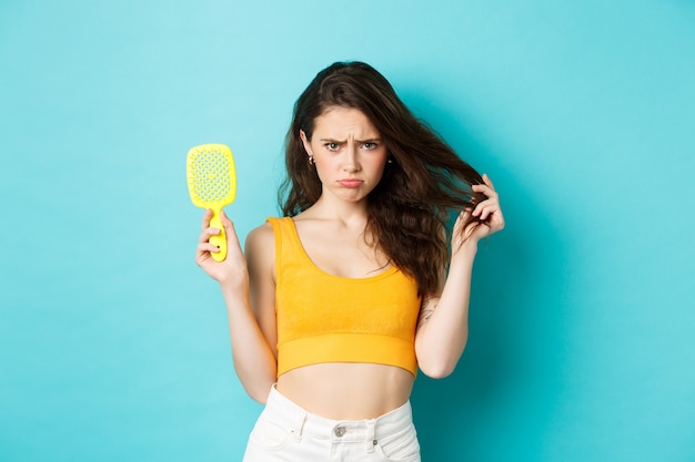 Mujer joven triste que muestra el cepillo con hebras, que tiene un problema de pérdida de cabello, necesita un tratamiento de belleza cosmético, de pie sobre fondo azul molesto