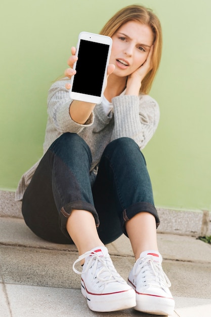 Foto gratuita mujer joven triste que se sienta contra la pared verde que muestra el teléfono móvil