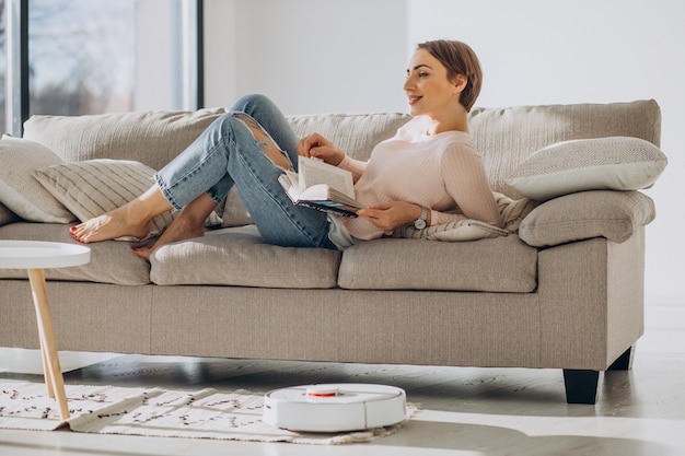 Mujer joven tumbado en el sofá y leyendo un libro mientras robot aspirador haciendo las tareas del hogar