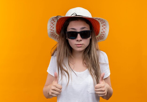 Mujer joven turista en camiseta blanca con sombrero de verano y gafas de sol negras que muestran los pulgares para arriba sobre la pared naranja