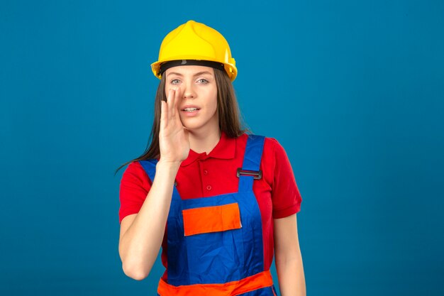 Mujer joven en uniforme de construcción y casco de seguridad amarillo con una mano cerca de la boca diciendo un secreto de pie sobre fondo azul.