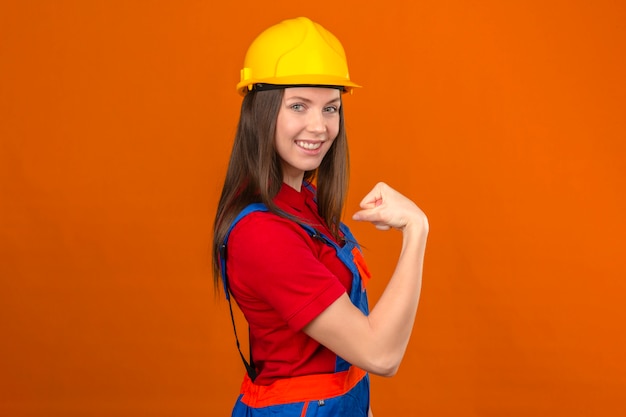Foto gratuita mujer joven en uniforme de construcción y casco de seguridad amarillo mostrando sus bíceps podemos hacerlo gesto sonriendo de pie sobre fondo naranja