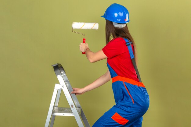 Mujer joven en uniforme de construcción y casco de seguridad azul en escalera con rodillo de pintura sobre fondo verde