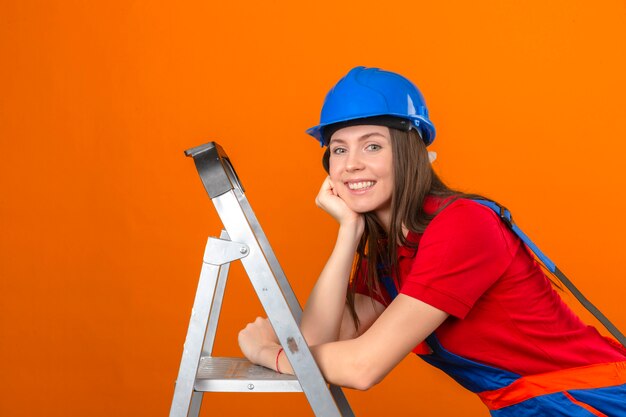 Mujer joven en uniforme de construcción y casco de seguridad azul en escalera con sonrisa en la cara sobre fondo naranja aislado