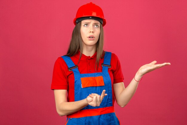 Mujer joven en uniforme de construcción y casco de seguridad rojo mirando hacia un lado contando y pensando de pie sobre fondo rosa oscuro